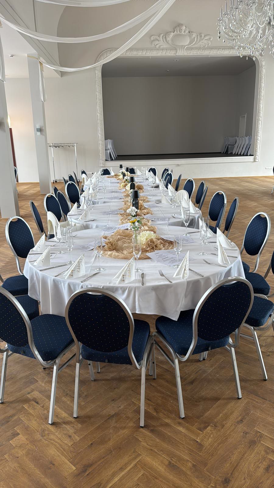 Lange Festtafel im Saal mit blauen Stühlen, dahinter die Bühne